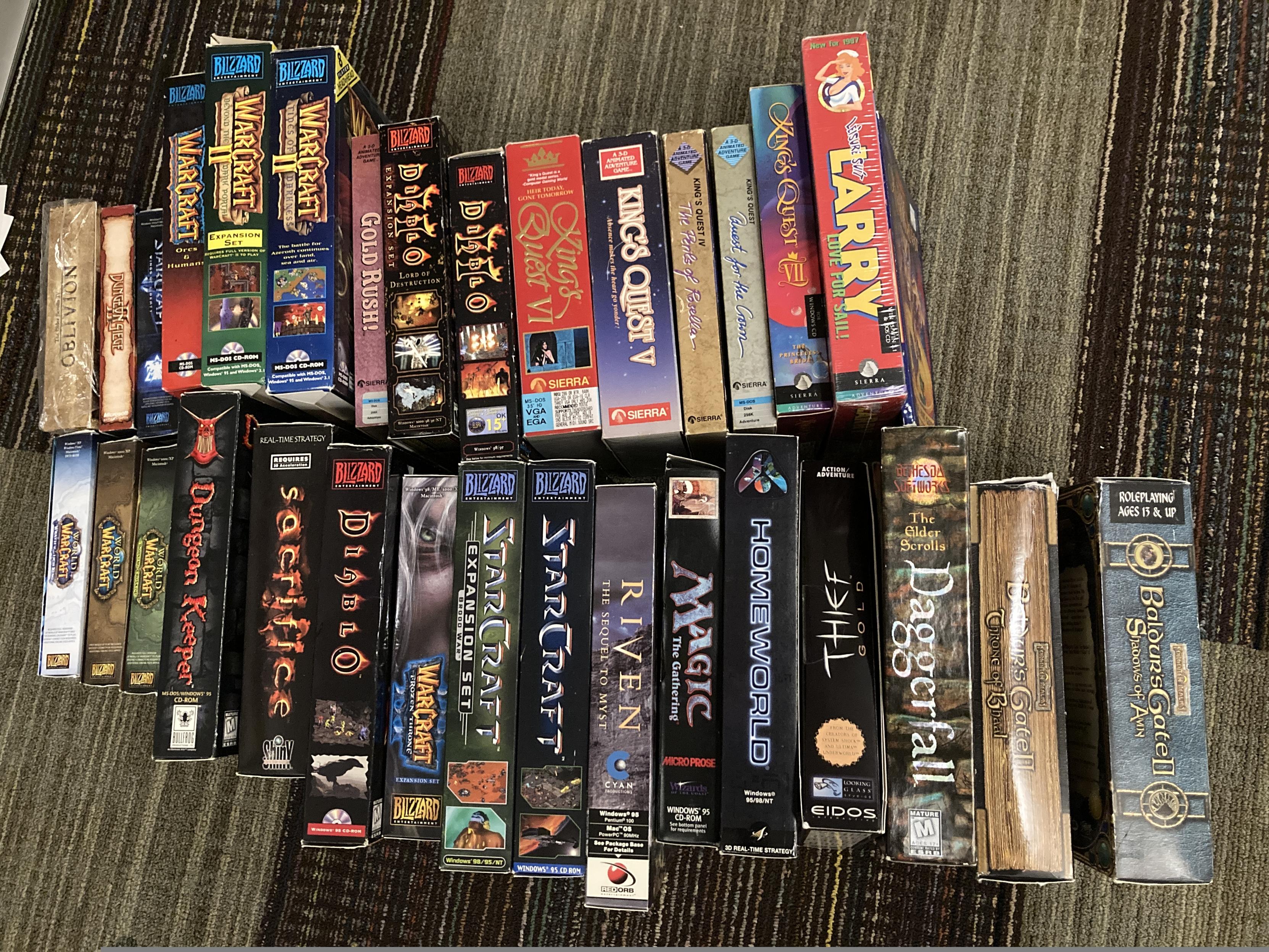 A bunch of big box PC games lined up on a floor. Left to right, top to bottom:

TES: Oblivion Collector's Edition
Dungeon Siege II
StarCraft II
WarCraft: Orcs & Humans
WarCraft II: Beyond the Dark Portal
WarCraft II: Tides of Darkness
Gold Rush!
Diablo II: Lord of Destruction
Diablo II
King's Quest VI: Heir Today, Gone Tomorrow
King's Quest V: Absence makes the heart go yonder
King's Quest IV: The Perils of Rosella
King's Quest I: Quest for the Crown
King's Quest VII
Leisure Suit Larry VI: Love for Sail!
World of Warcraft: Wrath of the Liche King
World of Warcraft
World of Warcraft: Burning Crusade
Dungeon Keeper
Sacrifice
Diablo
WarCraft III: Frozen Throne Expansion
StarCraft: Brood War
StarCraft
Riven
Magic: The Gathering
Homeworld
Thief Gold
Daggerfall
Baldur's Gate II: Throne of Bhaal
Baldur's Gate II: Shadows of Amn
